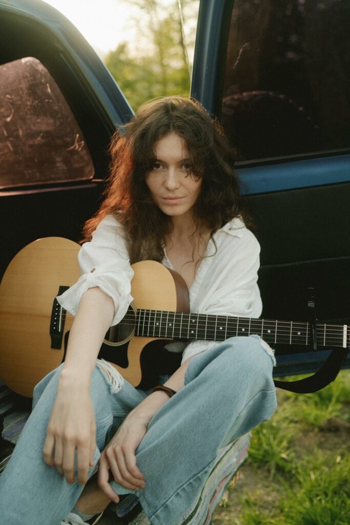 Jonge vrouw met gitaar zittend buiten bij een auto in de natuur.